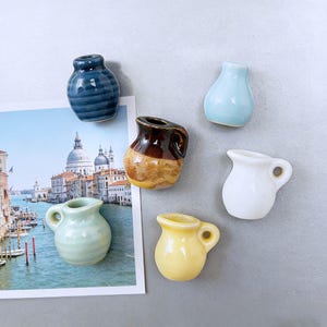 Puede incluir: Una colección de jarras y jarrones de cerámica en miniatura en varios colores, incluyendo azul, amarillo, blanco y verde. Algunos tienen asas, otros tienen superficies texturizadas. Una postal de una ciudad europea es parcialmente visible.