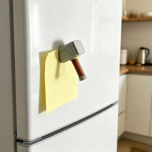 Könnte beinhalten: Ein hammerförmiger Magnet in Hellgrau mit braunem Griff, der auf der Seite ein Triquetra-Symbol aufweist. Der Magnet ist an einer weißen Kühlschranktür befestigt und hält einen einzelnen quadratischen gelben Zettel an der glatten Oberfläche.
