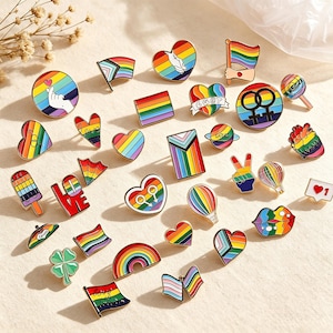 May include: A collection of metal enamel pins featuring various rainbow-themed designs, including hearts, flags, hands making gestures, hot air balloons, lips, and symbols. The pins display vibrant colours and shapes, including a heart with the text love.