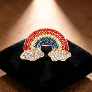 May include: A gold-toned brooch in the shape of a rainbow with two cloud accents. The rainbow is made of red, orange, yellow, green, and blue gemstones. The clouds are made of clear gemstones. The brooch is set on a black surface.