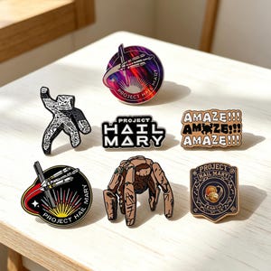 May include: A collection of six enamel pins featuring designs inspired by Project Hail Mary. The pins include a circular space mission emblem, a black and white figure, a logo, a text-based pin reading AMAZE!!!, a ship graphic, and a creature.