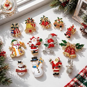 Pode incluir: Uma coleção de doze broches de metal com tema natalino, apresentando designs esmaltados de Papai Noel, bonecos de neve e sinos. Os alfinetes são decorados com esmalte colorido, strass transparentes e detalhes em metal dourado.