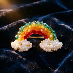 May include: A colorful rainbow brooch with a gold-tone pin. The rainbow features layers of red, orange, yellow, green, and clear crystals. White pearl-like beads form the clouds at each end of the rainbow.