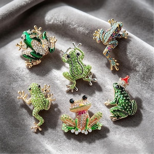 Puede incluir: Seis broches de rana enjoyados en varias poses. Los broches presentan cristales verdes, dorados y coloridos. Una rana lleva un gorro de Papá Noel. Los broches están colocados sobre una superficie gris texturizada.