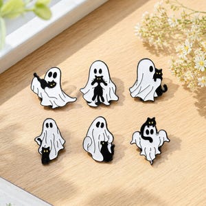May include: A set of six enamel pins featuring white ghosts with black cats. Each pin depicts a different interaction between a ghost and a black cat with yellow eyes. The designs include cats being held, sitting on shoulders, or peeking out from under the ghost sheets, all rendered in a high-contrast black and white style.