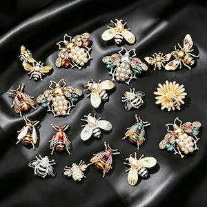 May include: A collection of various bee-shaped brooches featuring gold-tone metal, faux pearls, and colorful enamel accents. The pins display diverse designs, including some with crystal embellishments, striped bodies, and one featuring a daisy flower.