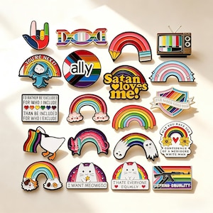 Könnte beinhalten: Eine Sammlung bunter Emaille-Anstecknadeln mit verschiedenen Motiven wie Regenbögen, Katzen, einer Gans, einem Fernseher und Texten wie "Satan loves me!", "I want meowgic", "I hate everyone equally", "Ally" und "Defend equality" auf hellem Grund.