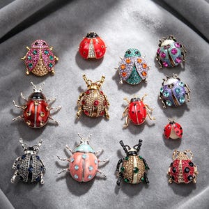 May include: A collection of decorative insect brooches in various colors and designs. The brooches feature ladybug and beetle shapes, adorned with colorful gemstones and enamel. The brooches are displayed on a gray surface.