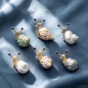 Pode incluir: Seis figuras decorativas de caracóis com corpos de metal dourado texturizado e conchas naturais em espiral. As conchas apresentam vários padrões e cores, incluindo castanho manchado, branco sólido e verde salpicado, dispostas em duas filas.