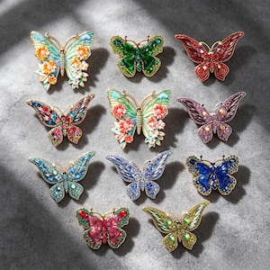 May include: A collection of eleven butterfly-shaped brooches arranged in a grid. Each brooch features intricate metalwork, vibrant enamel colors, and embedded rhinestones in various shades including red, blue, green, purple, and orange floral patterns.