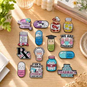Op de afbeelding: Een verzameling emaille pins met apotheek- en medicatiethema's, waaronder pillencapsules, medicijnflesjes en teksten zoals 'We Are Pharmily' en 'Eat Sleep Pharmacy Repeat' in diverse kleuren op een houten ondergrond.