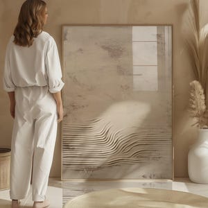 May include: A large framed abstract art piece with a neutral color palette of beige and brown tones. The artwork features a textured, layered design with horizontal lines. A woman in a white jumpsuit stands near the artwork. A white vase with dried plants is also visible.