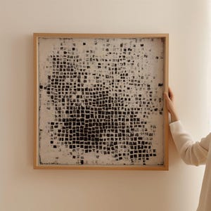 May include: Abstract artwork in a natural wood frame. The piece features a grid-like pattern of black and white squares, creating a textured appearance. The artwork is displayed on a neutral-colored wall.