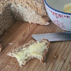 Puede incluir: Una rebanada de pan moreno con mantequilla, parcialmente comida, sobre una tabla de cortar de madera. Un pan entero está en el fondo, junto con un cuenco de mantequilla y un cuchillo. El pan tiene un aspecto rústico.