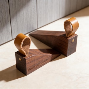 Puede incluir: Dos topes de puerta de madera marrón oscuro con forma triangular y un asa de bucle de cuero marrón claro. El texto "HOLD THE DOOR" está impreso en el lateral. Un pequeño remache dorado asegura el asa de cuero a la madera.
