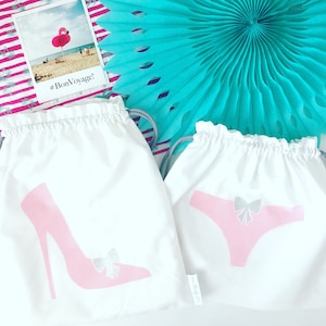 May include: Two white drawstring bags with different designs. One bag has a pink high heel shoe with a silver bow. The other bag has a pink bikini bottom with a silver bow. The text "#BonVoyage!" is visible on the bag with the high heel shoe.