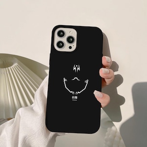 Puede incluir: Funda de teléfono negra con un diseño gráfico blanco. La funda presenta un diseño de rostro estilizado con texto japonés debajo. La funda del teléfono se muestra en una mano.