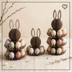 Puede incluir: Tres soportes de madera para huevos de Pascua con forma de conejos. Cada conejo tiene una bandeja escalonada llena de huevos de Pascua de colores pastel. Los soportes son marrones y los huevos están decorados con varios diseños.