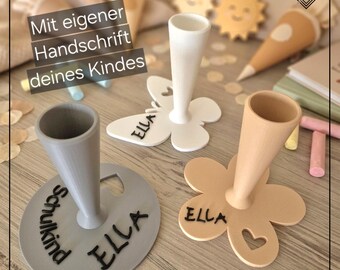 May include: Three cone-shaped holders with flat bases in white, gray, and beige. The white base is butterfly-shaped, the gray is circular, and the beige is flower-shaped. Each features the name ELLA and the text Mit eigener Handschrift deines Kindes.