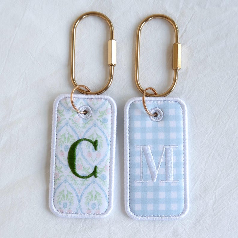 Op de afbeelding: Twee rechthoekige sleutelhangers met goudkleurige hardware. E&eacute;n sleutelhanger heeft een groene "C" op een bloemenpatroon, de andere een witte "M" op een blauw geruit patroon. Beide hebben wit gestikte randen.