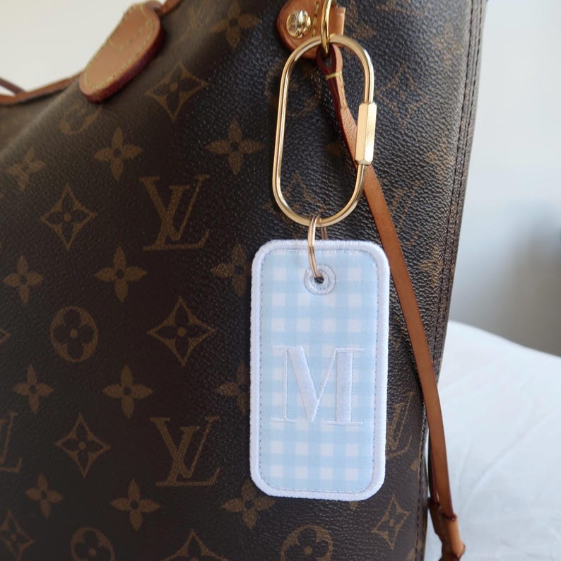 Op de afbeelding: Een lichtblauwe gingham bagagelabel met een wit geborduurde letter "M" is bevestigd aan een goudkleurige sluiting. Het label hangt aan een bruin lederen riem die is bevestigd aan een bruine en tan handtas met patroon.