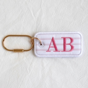 Op de afbeelding: Een rechthoekige wit en roze gestreepte sleutelhanger met de geborduurde letters "AB" in roze. De sleutelhanger heeft een goudkleurige metalen karabijnhaak om aan sleutels of tassen te bevestigen.