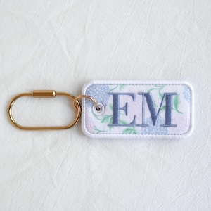 Op de afbeelding: Een rechthoekige sleutelhanger met de geborduurde letters "EM" in blauw. De sleutelhanger heeft een bloemenpatroon in blauwe en roze tinten. Hij is bevestigd aan een goudkleurige karabijnhaak.