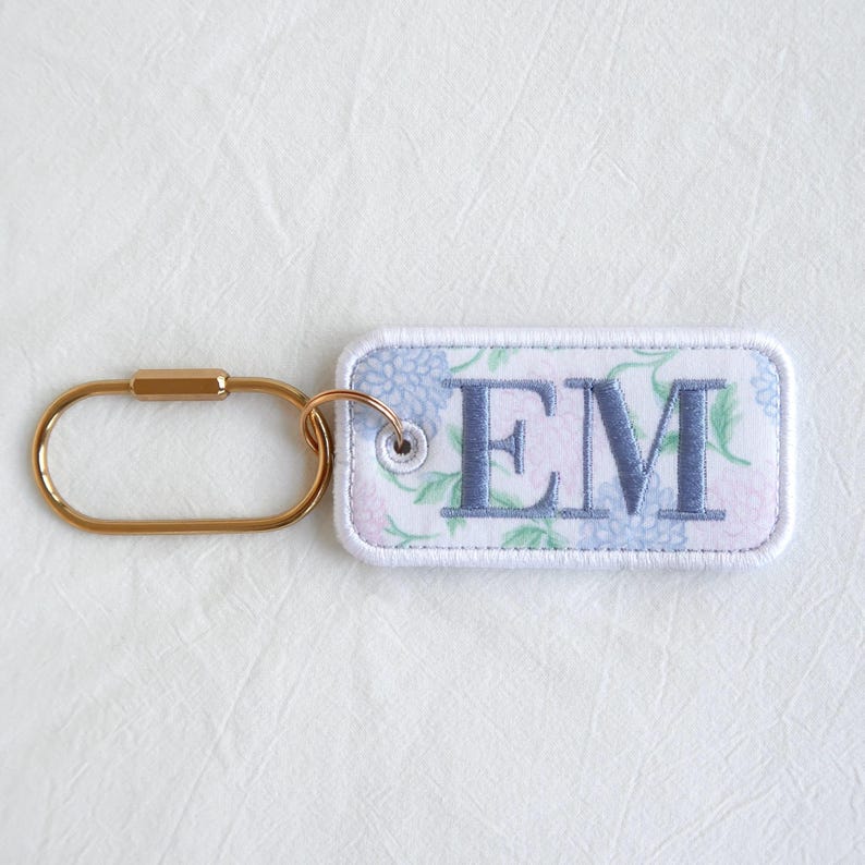 Op de afbeelding: Een rechthoekige sleutelhanger met de geborduurde letters "EM" in blauw. De sleutelhanger heeft een bloemenpatroon in blauwe en roze tinten. Hij is bevestigd aan een goudkleurige karabijnhaak.