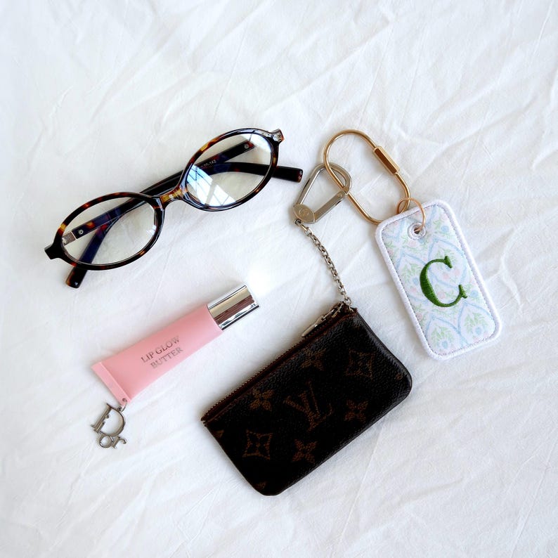 Op de afbeelding: Een verzameling accessoires op een witte ondergrond. Inclusief een bril met schildpadmotief, een roze lipgloss tube, een kleine bruine portemonnee en een sleutelhanger met een groen geborduurde letter "C".