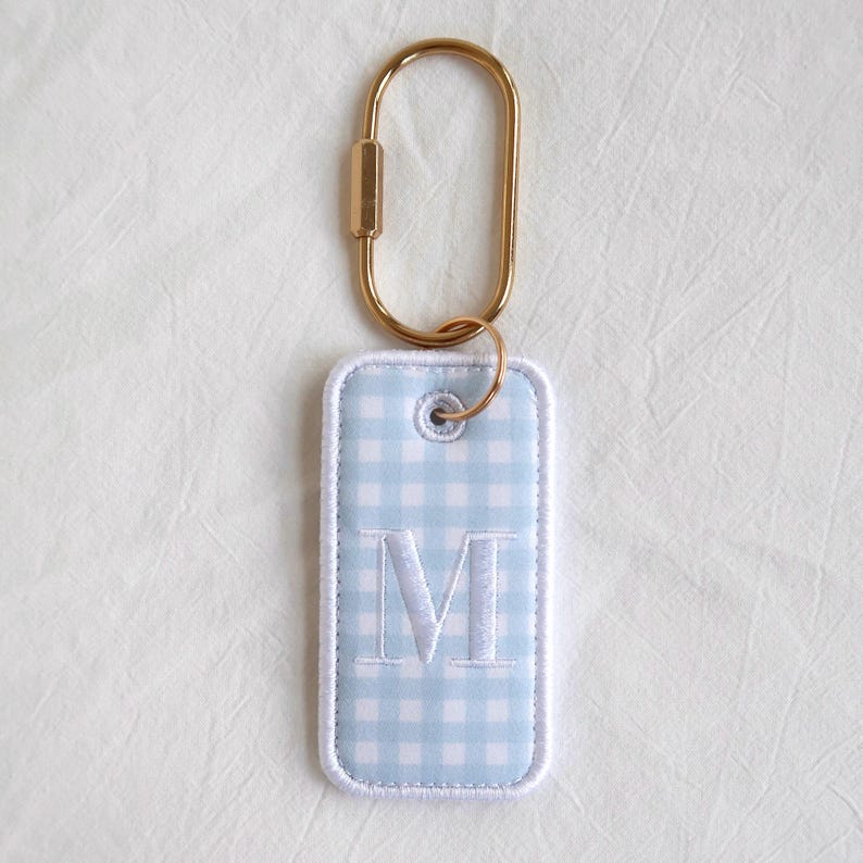 Op de afbeelding: Een rechthoekige sleutelhanger met een lichtblauw en wit ruitpatroon en een wit geborduurde letter "M". De tag is bevestigd aan een goudkleurige karabijnhaak. De tag heeft een witte rand.