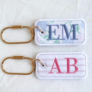 Op de afbeelding: Twee rechthoekige sleutelhangers met goudkleurige metalen sluitingen. E&eacute;n sleutelhanger heeft een bloemenpatroon met blauw geborduurde letters "EM". De andere heeft roze gestreepte stof met roze geborduurde letters "AB".
