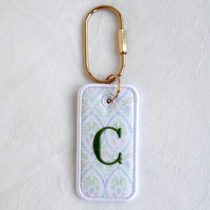 Op de afbeelding: Een rechthoekige sleutelhanger met een goudkleurige karabijnhaak. De sleutelhanger heeft een witte rand en een patroonstof met blauwe, groene en roze bloemmotieven. Een groene geborduurde letter "C" is gecentreerd op de stof.