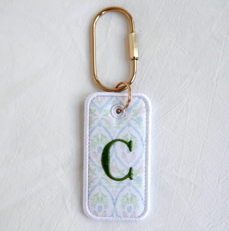 Op de afbeelding: Een rechthoekige sleutelhanger met een goudkleurige karabijnhaak. De sleutelhanger heeft een witte rand en een patroonstof met blauwe, groene en roze bloemmotieven. Een groene geborduurde letter "C" is gecentreerd op de stof.