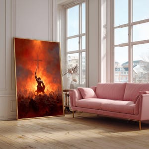 May include: A framed artwork depicts a warrior holding a sword and a cross aloft against a fiery backdrop. The artwork is in a gold frame and leans against a white wall. A pink sofa and a window are in the background.