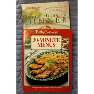 May include: Two Betty Crocker cookbooks are stacked. The top book is titled "Come Home to Dinner", and the bottom book is "30-Minute Menus". The bottom book features a photo of a meal on a green plate. The books are on a blue surface.