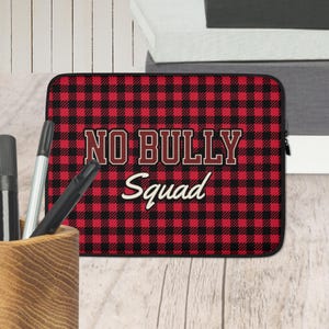 May include: Laptop sleeve with a red and black buffalo plaid pattern. The text "NO BULLY Squad" is displayed in a bold, outlined font. The sleeve is designed to protect a laptop and has a zipper closure.