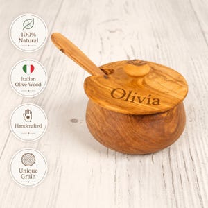 Può includere: Una ciotola in legno fatta a mano con coperchio e cucchiaio, realizzata in legno di ulivo italiano. Il coperchio è inciso con il nome "Olivia". La ciotola è rotonda con una finitura naturale a venatura del legno. L'immagine include anche etichette circolari con testo.