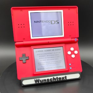 Könnte beinhalten: Ein rotes Nintendo DS-Handheld-Spielgerät, das geöffnet ist, um zwei Bildschirme anzuzeigen. Der obere Bildschirm zeigt das Nintendo DS-Logo. Der untere Bildschirm zeigt Text an. Ein schwarz-weißer Ständer mit dem Wort "Wunschtext" befindet sich vor der Konsole.
