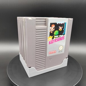 Könnte beinhalten: Ein Stapel Retro-Spielkassetten für das Nintendo Entertainment System (NES) mit dem Spiel "Kid Icarus" im Vordergrund. Die Kassetten sind grau mit einem weißen Etikett mit Spielgrafiken und dem Nintendo-Logo. Der Stapel wird in einem weißen Halter gehalten.