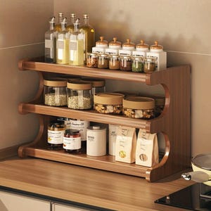 Puede incluir: Un especiero de madera de tres niveles lleno de varios frascos, botellas y recipientes de vidrio. El estante es de madera marrón oscuro y se encuentra sobre una encimera. Los frascos contienen especias, granos y líquidos.