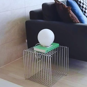 May include: A modern side table with a chrome wireframe design. A white globe lamp sits atop a green book on the table. The table is next to a black leather sofa with decorative pillows.