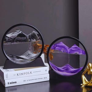 May include: Two circular sand art pieces with black frames and bases. One features black and gold sand, the other purple sand, creating mountain scenes. The pieces sit on a stack of books with the text "The Kinfolk" and "Union".