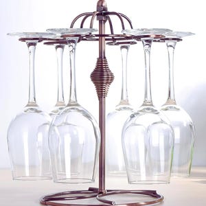 May include: A bronze-colored metal wine glass rack with a circular base and a central support. The rack holds six clear wine glasses, suspended upside down. The design features decorative spiral detailing and a circular top for the glass stems.