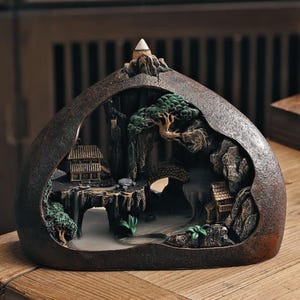 May include: A decorative incense burner shaped like a mountain landscape. The brown resin sculpture features miniature houses, trees, a bridge, and a waterfall. A cone-shaped incense holder sits atop the mountain, with smoke rising from the base.
