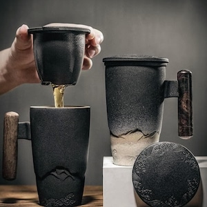 Könnte beinhalten: Schwarze Keramik-Teetasse mit Holzgriff und passendem Deckel. Die Tasse hat ein Bergdesign und einen Tee-Infuser. Der Tee wird in die Tasse gegossen. Die Tasse ist etwa 13 cm hoch.