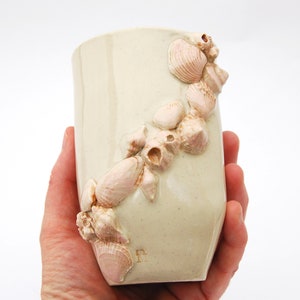 May include: A white ceramic mug with a raised design of pink seashells. The mug is being held in a hand.