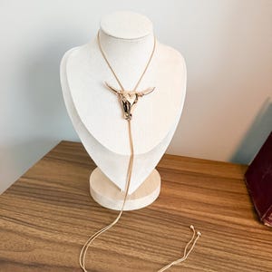 May include: A rose gold-toned bolo tie necklace featuring a polished metal steer skull pendant. The long, slender chain passes through the back of the steer head, ending in two small metal beads at the bottom of the dangling cord strands.
