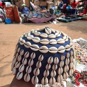 May include: A deep blue dome-shaped cap decorated with rows of white cowrie shells. The top features concentric circles of shells, while the sides are adorned with vertical strands of shells arranged in a repeating pattern around the entire base.
