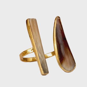 Könnte beinhalten: Ein goldfarbener Ring mit zwei markanten Elementen: ein vertikales, rechteckiges Element und ein gebogenes, tropfenförmiges Element. Beide haben eine hellbraune und cremefarbene Farbe mit goldenen Rändern. Das Band des Rings ist ebenfalls goldfarben.