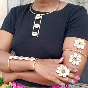 May include: A gold-toned jewelry set featuring a necklace, bracelet, arm cuff, and ring. The necklace has three shell pendants. The bracelet has six shell pendants. The arm cuff and ring have flower-shaped designs. The jewelry is worn over a black top.